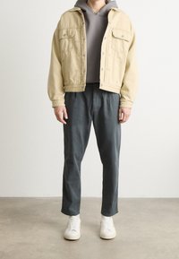 Beige denim jacket yli harmaa huppari, yhdistettynä löysiin tummanharmaisiin housuihin ja valkoisiin lenkkikengiin. Ominaisuuksia ovat nappisuljenta ja sivutaskut.