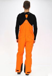 Oranje waterdichte bibluren met verstelbare schouderbanden, voorzien van zwarte accenten bij de enkels en zakken. Glad textuur, duurzaam ontwerp.