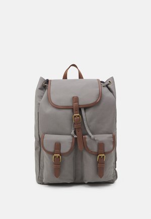 Sac à dos en tissu gris avec des sangles à boucle en cuir marron sur le rabat principal et deux poches avant, ainsi qu'une poignée de transport supérieure.