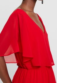 Robe rouge avec un décolleté en V plongeant, des manches flottantes transparentes et un tissu texturé avec une taille élastique pour une silhouette ajustée.