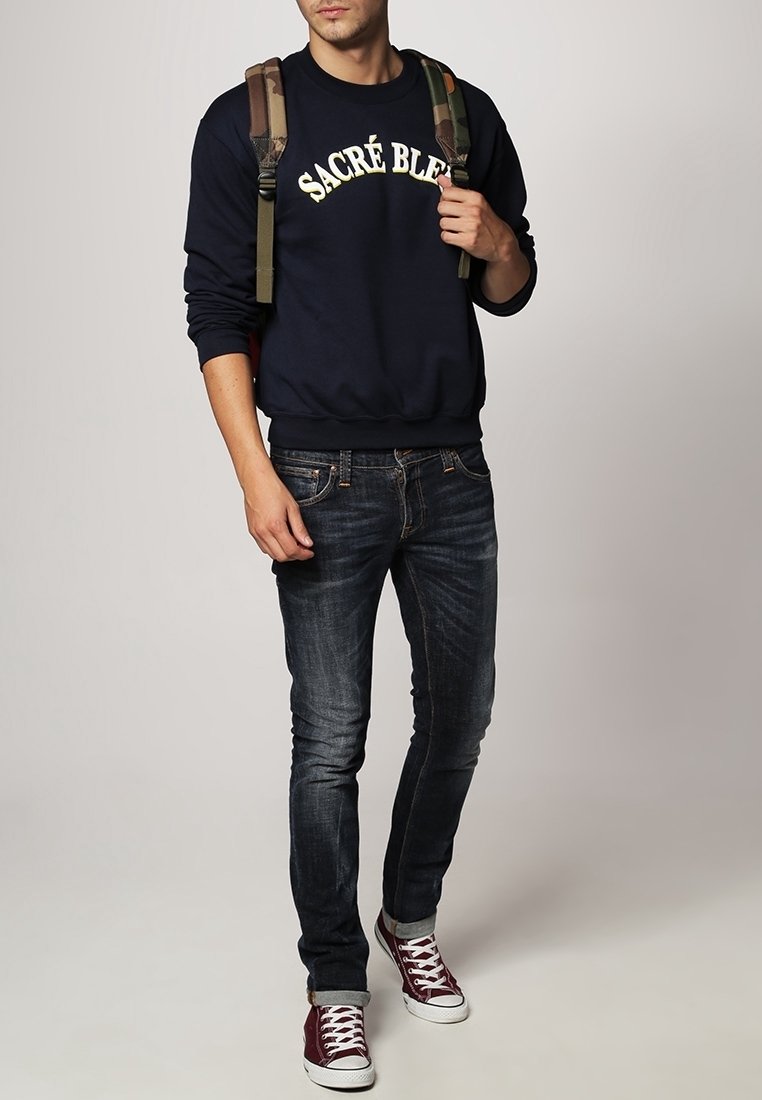 Giovane uomo che indossa una felpa blu navy con la scritta "SACRÉ BLEU", jeans scuri, sneakers rosse e ha uno zaino mimetico appoggiato sulle spalle.