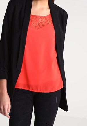 Vrouw draagt een fel koraalroze mouwloos top met kantdetail onder een zwarte open blazer en zwarte broek.