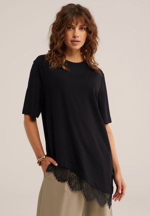 Vrouw met krullend bruin haar, gekleed in een loszittende zwarte top met korte mouwen en een kanten zoom, over een beige rok, staand met één hand in haar zak.