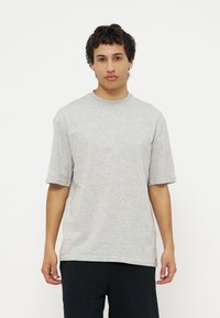 Νεαρός άνδρας με κοντά σγουρά μαλλιά, φορώντας ένα άνετο ανοιχτό γκρι κοντομάνικο πουκάμισο και μαύρο παντελόνι, στέκεται μπροστά σε ένα απλό λευκό φόντο.