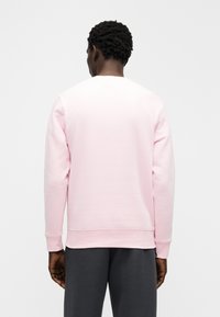 Sweatshirt rosa claro com mangas compridas, gola redonda e punhos canelados. O tecido tem uma textura suave, combinado com calças cinzentas escuras.
