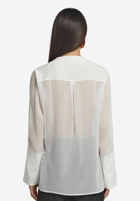 Blusa bianca a maniche lunghe con retro trasparente, polsini con bottoni, colletto arrotondato e una texture liscia, caratterizzata da un design a strati.