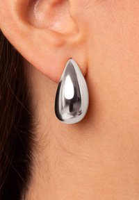 Boucle d'oreille en argent en forme de goutte ; surface lisse et réfléchissante ; finition métallique polie ; attachée au lobe de l'oreille ; sans embellissements visibles.