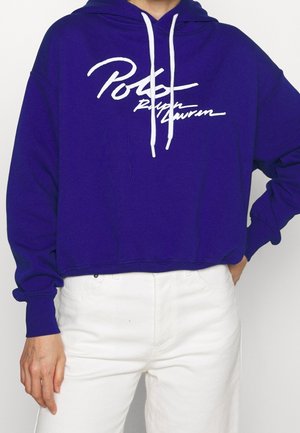 Persoon die een paarse cropped hoodie draagt met witte trekkoorden en witte sierlijke tekst, gecombineerd met witte broeken met een hoge taille.