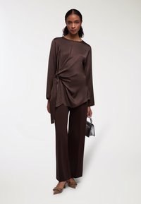 Top in satin marrone con dettaglio incrociato, abbinato a pantaloni a palazzo coordinati. Completato con una piccola borsa nera e scarpe beige.