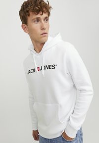 Hvit bomullshoodie med frontlomme i kenguru-stil, justerbar snor til hetta, og svart "JACK & JONES" logo over brystet.