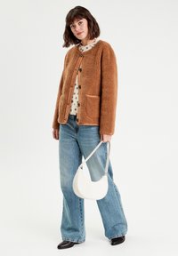 Veste en polaire texturée marron avec des boutons noirs, deux poches avant, portée sur un haut crème à pois marron, associée à un jean bleu clair.