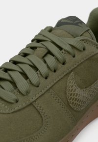 Maslinastozelena Nike tenisica s teksturiranim Swoosh logom, odgovarajućim vezicama, nubuk gornjim dijelom i gumenim potplatom, prikazana iz blizine s nagibom.