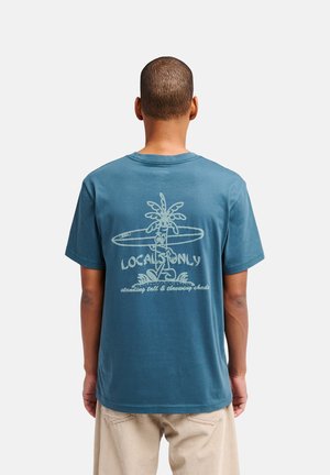 Homme portant un t-shirt bleu avec un dessin de palmier et de planche de surf ainsi que le texte « LOCALS ONLY standing tall & throwing shade » dans le dos.