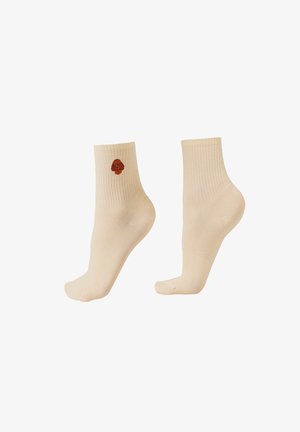 Béžové ponožky do kotníků, jedna z nich má u manžety vyšívaný malý hnědý psí obličej, žebrovaná textura.