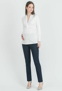 Gravidtröja i vitt jersey med tvinnat frontdetalj, matchad med marinblå figurnära byxor. Modellen står med en hand på magen.