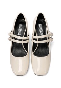 Scarpe Mary Jane in pelle verniciata beige con doppie cinghie, punta quadrata e suole nere, dotate di una texture liscia e lucida.