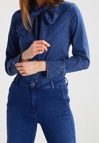 Denimskjorta i mörkblått med en stor fluga vid kragen. Har långa ärmar och en figurnära design. Matcherade med blå jeans.