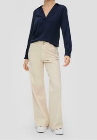 Dunkelblaue Bluse mit langen Ärmeln, V-Ausschnitt und subtilen Falten; kombiniert mit cremefarbenen, weiten Hosen und weißen Sneakers für eine legere Kleidung.