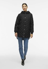 Črna prešita jakna s kapuco, gumbi na pritiskanje in stranskimi žepi. V kombinaciji z modrimi naravnimi hlačami in črnimi pikastimi gležnarji.