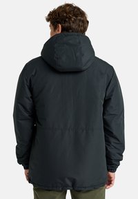 Hombre con una chaqueta de invierno negra con capucha visto desde atrás sobre un fondo claro y liso.