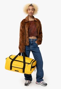 Bolsa de deporte amarilla con correas y detalles de cremallera en negro, que presenta una amplia apertura, material resistente y forma rectangular.