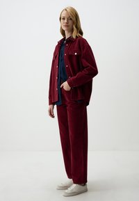 Bordeaux corduroyjas en -broek met een geknoopte marineblauwe blouse eronder. Model draagt witte sneakers en staat tegen een neutrale achtergrond.