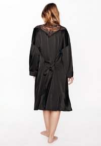 Robe en satin noir à manches longues, avec un col orné de dentelle, une fermeture à nouer à la taille, et une coupe fluide arrivant au genou.