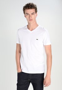 Balta medvilninė marškinėlių su V formos iškirpte, trumpomis rankovėmis ir mažuoju žaliu "Lacoste" logotipu kairėje krūtinės pusėje. Lygus paviršius, prigludusi forma.