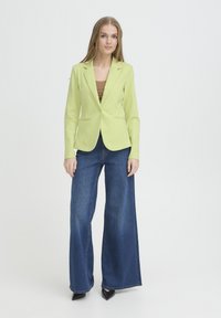 Mujer con blazer verde claro, top marrón, vaqueros de pierna ancha azules y zapatos negros puntiagudos, de pie contra fondo blanco.