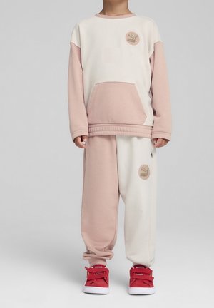 Lapsi, joka pukeutunut beigen ja valkoisen sävyiseen Puma-vapaa-ajanasuunsa ja punaisiin lenkkareihin, seisoo yksinkertaisen harmaan taustan edessä.