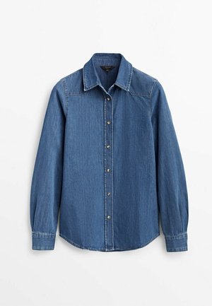 Chemise en denim bleu à manches longues avec boutons-pression devant, col pointu et poignets boutonnés sur fond blanc uni.