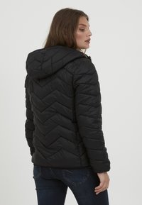 Doudoune noire avec un motif matelassé en chevrons, une capuche et une fermeture éclair à l'avant. Le design présente des poignets élastiques et une silhouette courte.