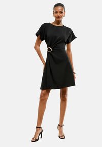 Vestido negro de manga corta que presenta una cintura ceñida con un acento circular dorado, tejido suave y una silueta relajada, que cae por encima de las rodillas.