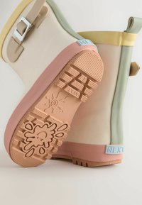 Stivali di gomma colorati con corpo beige, dettagli verdi, suola rosa e un motivo di grip testurizzato. Cinghia con dettaglio in fibbia metallica.