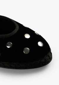 Zapatilla de terciopelo negro con punta redondeada, adornada con tachuelas metálicas dispuestas en un patrón. Incluye una suela texturizada y un ribete elástico en el tobillo.