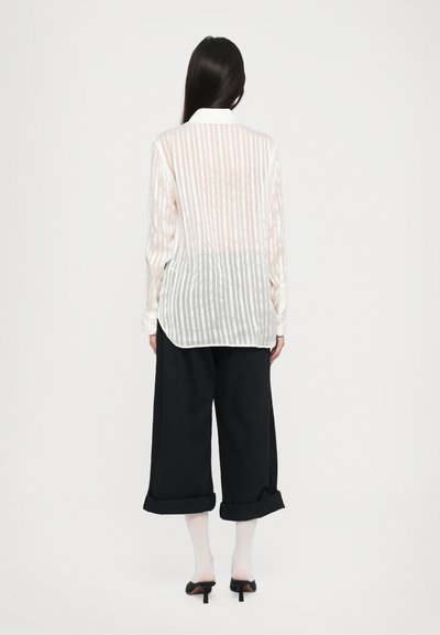 Przezroczysta, długotaliowa koszula w jasnoróżowe i białe paski, z tylnym karczkiem i zaokrąglonym hemem. Zestawiona z szerokimi czarnymi spodniami i szpilkami.