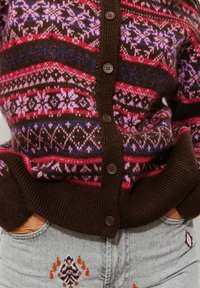 Cardigan tricoté marron avec des motifs géométriques roses, rouges et bleus, présentant cinq boutons marron et des poignets côtelés. Porté sur un jean gris avec des broderies.