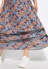 Frau hält einen floral gemusterten, plissierten Rock mit durchsichtigen schwarzen Netzstreifen, trägt weiße klobige Sneakers und steht vor einem weißen Hintergrund.