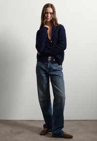 Marineblaues Strickcardigan mit tiefem V-Ausschnitt und Knopfverschluss, kombiniert mit hellblauen, weit geschnittenen Jeans und braunen flachen Schuhen.