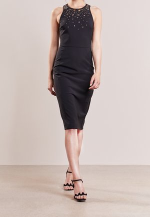 Zwarte mouwloze jurk met een nauwsluitend silhouet, versierd met kleine metalen sterren bij de halslijn, gecombineerd met zwarte sandalen met bandjes.