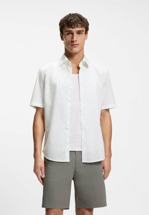 Joven con camisa blanca de manga corta abotonada sobre una camiseta blanca sin mangas y pantalones cortos grises, de pie contra un fondo liso.