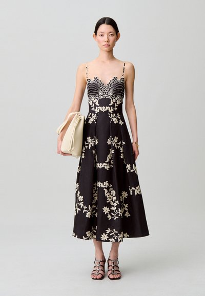 Abito midi nero con ricamo floreale color crema, spalline sottili, corpetto aderente con dettaglio smerlato, gonna fluente. Modella tiene una borsa beige.
