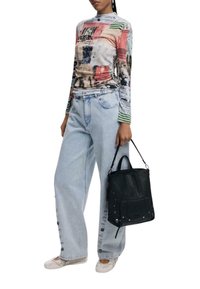 Donna che indossa una camicia a maniche lunghe colorata, jeans larghi chiari con bottoni laterali, scarpe bianche e tiene una borsa in pelle nera per il manico.