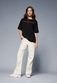 Camiseta negra oversize con logo rosa, pantalones de pierna ancha color crema y sneakers blancos con detalles rosa, llevados por un modelo de pie contra un fondo gris.