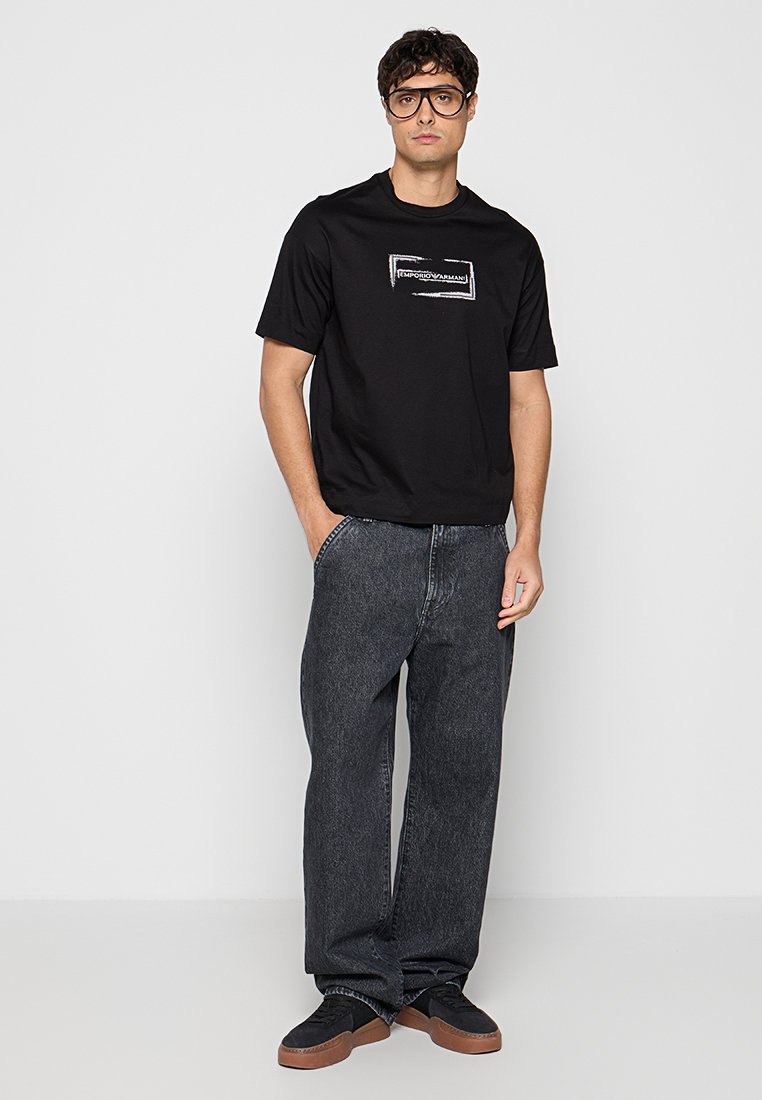 Hombre con camiseta negra de Emporio Armani, vaqueros holgados de color gris oscuro, zapatillas negras y gafas grandes negras, de pie contra un fondo gris claro.
