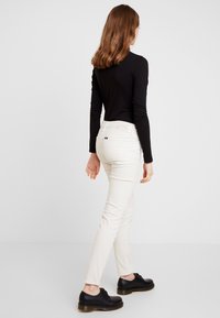 Kvinna med axellångt brunt hår som bär en svart långärmad tröja, vita jeans och svarta skor, stående och tittande åt sidan.