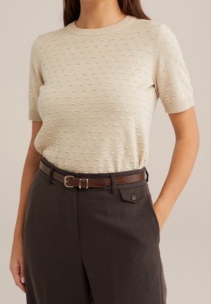 Femme portant un pull beige texturé à manches courtes, rentré dans un pantalon taille haute marron foncé avec une ceinture en cuir marron.