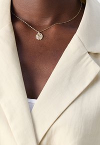 Collier avec pendentif rond texturé doré sur peau foncée, porté avec un blazer crème sur un top blanc.