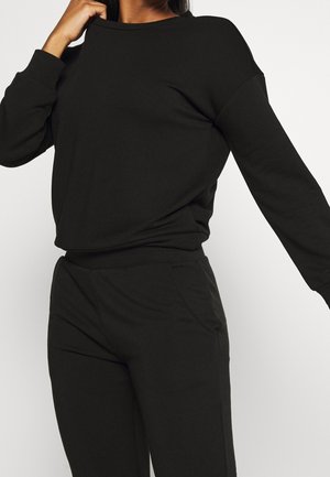 Frau trägt ein einfarbiges schwarzes Langarmshirt und passende schwarze Hose, mit einem Arm angewinkelt und dem anderen ausgestreckt.