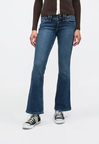 Džínsy z modrého denimu v strednom odtieni modrej, s vysokým pásom, piatimi vreckami a kontrastnými švami. Kombinované s čiernymi teniskami.